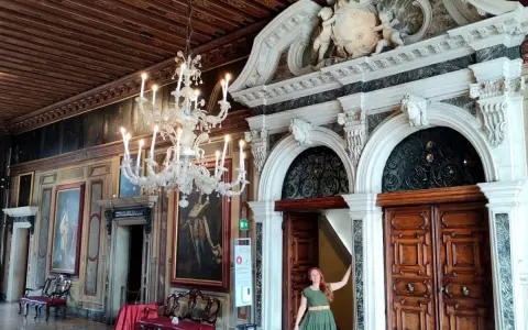 Una donna in un abito verde posa davanti a un portale decorato in una casa museo a Venezia, con un lampadario di cristallo e dipinti alle pareti.