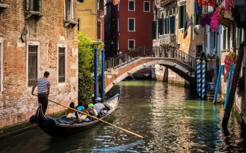 Gondola con turisti naviga in un canale, sotto un ponte, scoprendo la Venezia segreta tra palazzi antichi e fiori.