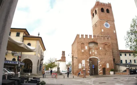 Piazza con torre medievale, portici e negozi. Ecco cosa vedere a Noale: la torre civica con orologio, il palazzo giallo e i tavolini all'aperto.
