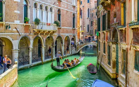 Un gondoliere a Venezia guida una gondola con turisti lungo un canale verde, tra palazzi storici e un ponte affollato.