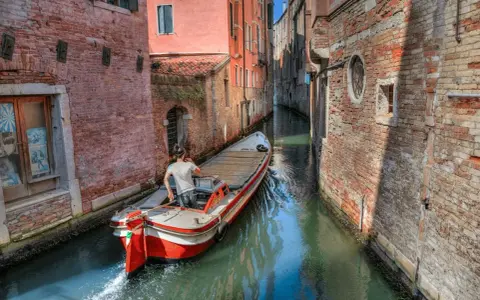 Un uomo guida una barca su un canale stretto a Venezia, circondato da edifici in mattoni. Quanti canali ci sono a Venezia? Molti, come questo!