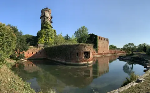 Antica torre e mura di mattoni circondate da un fossato, un angolo suggestivo di Venezia e dintorni, con vegetazione rigogliosa.