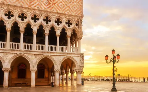 Palazzi storici di Venezia all'alba, con il sole che illumina la piazza e le architetture gotiche.