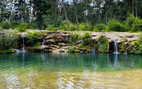 Cascate naturali nella valle di Cadore, con acqua cristallina che scende tra rocce e vegetazione rigogliosa, circondate da un bosco di pini.