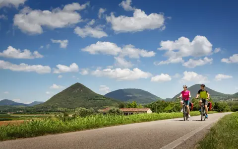 Coppia in bicicletta lungo la ciclabile dei Colli Euganei, tra verdi colline e cielo azzurro con nuvole bianche.