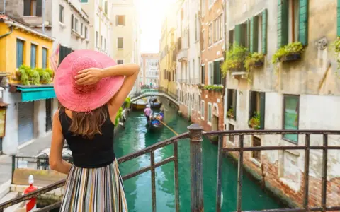 Donna con cappello rosa ammira un canale a Venezia, con gondole e palazzi storici. Un'esperienza da vivere a piedi, tra Santa Lucia e Piazza San Marco.