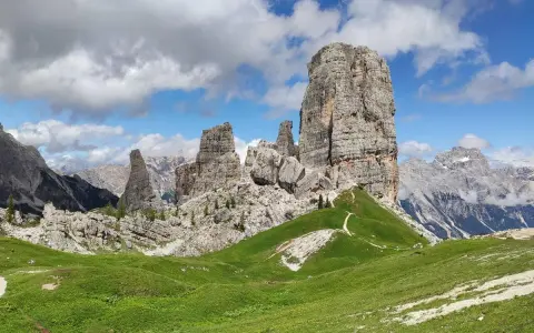 Sentiero che serpeggia tra verdi prati verso le maestose Cinque Torri Dolomiti, sotto un cielo azzurro con nuvole bianche.