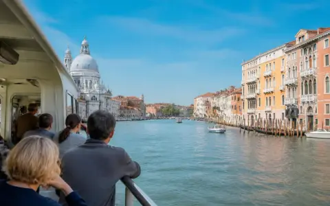 Gente sul vaporetto ammira il Canal Grande, con palazzi colorati e la Basilica di Santa Maria della Salute. Un viaggio tra Murano e Burano.