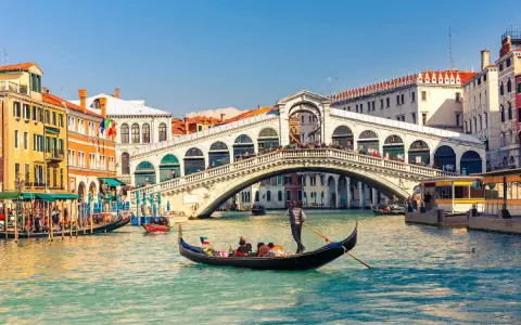 Una gondola solca il Canal Grande a Venezia, passando sotto il Ponte di Rialto, con edifici storici e altre imbarcazioni sullo sfondo.