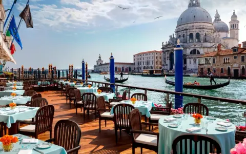 Tavoli apparecchiati con tovaglie azzurre e fiori arancioni, affacciati sul Canal Grande, con gondole e la Basilica di Santa Maria della Salute. Un'ambientazione da sogno per i migliori ristoranti di Venezia.