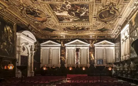 Sala capitolare Scuola Grande di San Rocco: soffitto dorato affrescato, pareti con dipinti e statue, sedie rosse disposte per un evento.