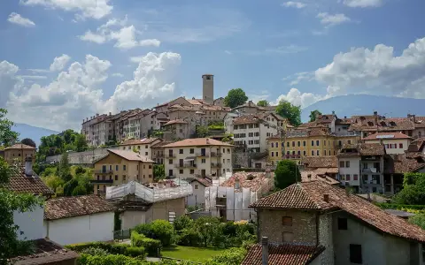 Panoramica di Feltre e dintorni, con case dai tetti rossi che si arrampicano sulla collina sotto un cielo azzurro e nuvoloso.