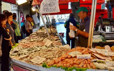 Mercato Venezia: bancarella di pesce fresco con gamberi, calamari e pesciolini. Venditore con cappellino verde.