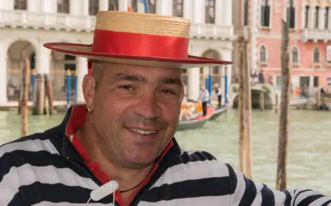 Un gondoliere sorridente, con il suo cappello iconico, pronto a navigare i canali di Venezia, la città che porta il soprannome di "La Serenissima".