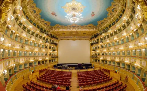 Interno sfarzoso del Teatro La Fenice di Venezia, con palchi dorati, soffitto affrescato e un grande schermo bianco.