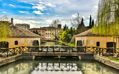 Ponte in legno su un canale con mulini ad acqua, case storiche e vegetazione rigogliosa, un'atmosfera serena che ricorda Portogruaro Caorle.