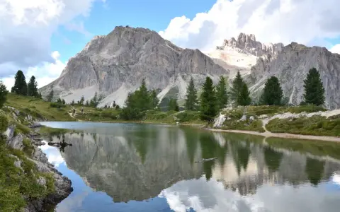 Riflessi perfetti delle montagne nel **lago di Limedes**, con sentiero e pini che incorniciano il paesaggio.