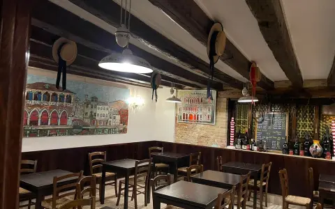 Interno di un **ristorante antico** con dipinti di **Venezia**, tavoli in legno e un calice di vino.