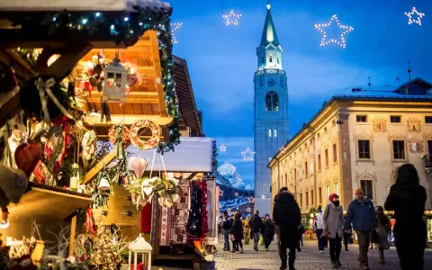 Mercatino di Natale a Cortina d'Ampezzo, con bancarelle addobbate, luci scintillanti e la torre del campanile che domina il cielo serale.