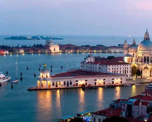 La **Punta della Dogana** a Venezia, con la Basilica di Santa Maria della Salute sullo sfondo, illuminata al crepuscolo. Barche e yacht ormeggiati nell'acqua calma.