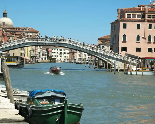 Un motoscafo naviga sotto uno dei ponti di Venezia, con edifici storici e gente che passeggia.