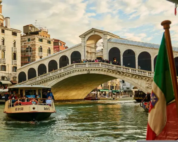 Un vaporetto naviga sul Canal Grande, passando sotto il Ponte di Rialto. L'Italia è rappresentata da una bandiera sventolante.