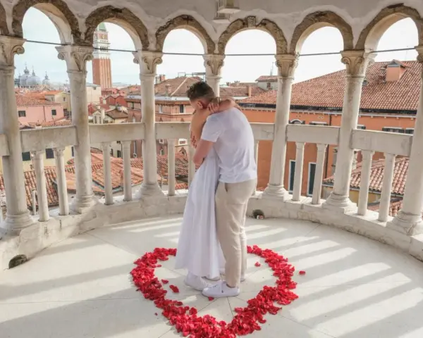 Un uomo abbraccia una donna su un balcone con vista su Venezia, circondati da petali di rosa a forma di cuore. Una perfetta proposta matrimonio Venezia.
