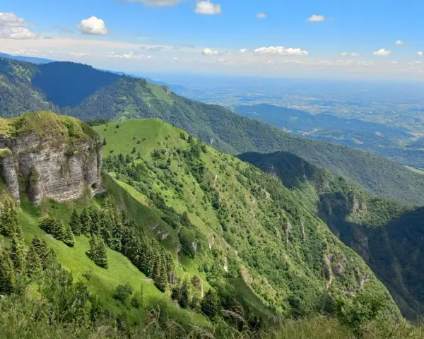 Panoramica mozzafiato dal Passo San Boldo, con verdi pendii montuosi che scendono verso una valle lontana sotto un cielo azzurro con nuvole bianche.