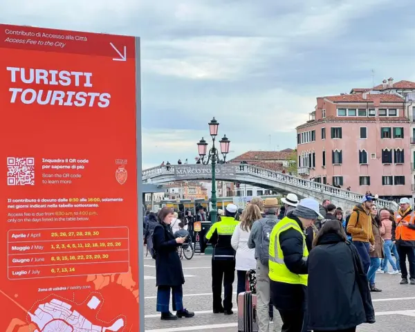 Cartello rosso a Venezia per il contributo accesso turisti, con date e QR code. Persone in fila sullo sfondo.
