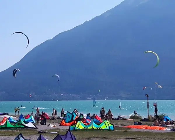 Coloratissima spiaggia sul lago di Santa Croce, con kitesurfisti che sfrecciano sull'acqua turchese e vele colorate che ondeggiano al vento.