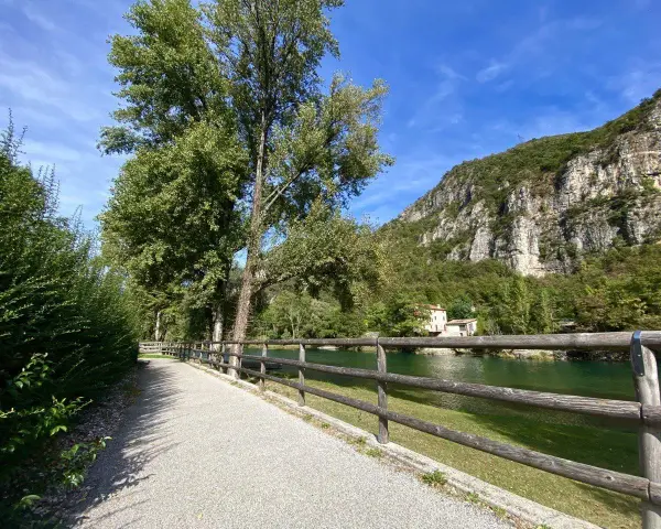 Sentiero ghiaioso lungo il fiume, con alberi verdi e una montagna rocciosa sullo sfondo. La **ciclabile del Brenta** invita a una passeggiata.