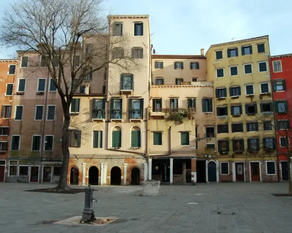 Edifici colorati nel ghetto ebraico di Venezia, con un albero spoglio e una fontana nella piazza lastricata.
