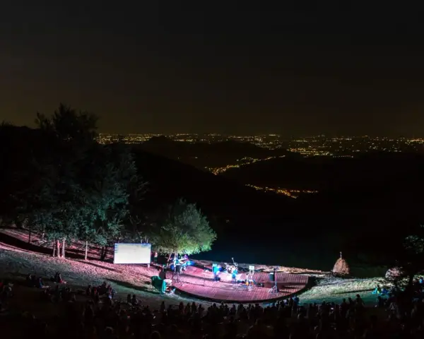 Concerto serale all'anfiteatro del Venda, con un pubblico seduto sull'erba e una band sul palco illuminato, sullo sfondo di un panorama notturno di luci cittadine.