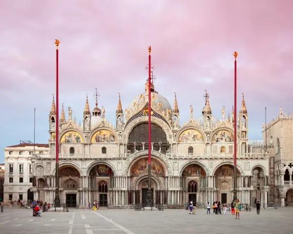 La maestosa basilica di San Marco a Venezia, con le sue cupole e mosaici dorati, si staglia sotto un cielo rosa al tramonto.