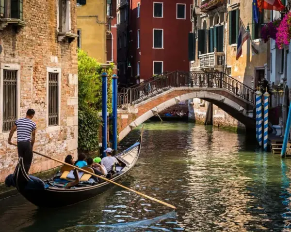 Gondola con turisti naviga in un canale, sotto un ponte, scoprendo la Venezia segreta tra palazzi antichi e fiori.