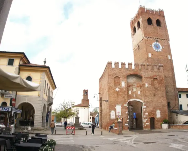 Piazza con torre medievale, portici e negozi. Ecco cosa vedere a Noale: la torre civica con orologio, il palazzo giallo e i tavolini all'aperto.