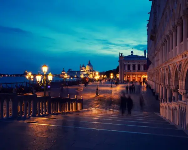 Venezia di notte, con le luci dei lampioni che illuminano la piazza e le sagome sfocate dei passanti.