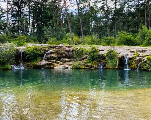 Cascate naturali nella valle di Cadore, con acqua cristallina che scende tra rocce e vegetazione rigogliosa, circondate da un bosco di pini.