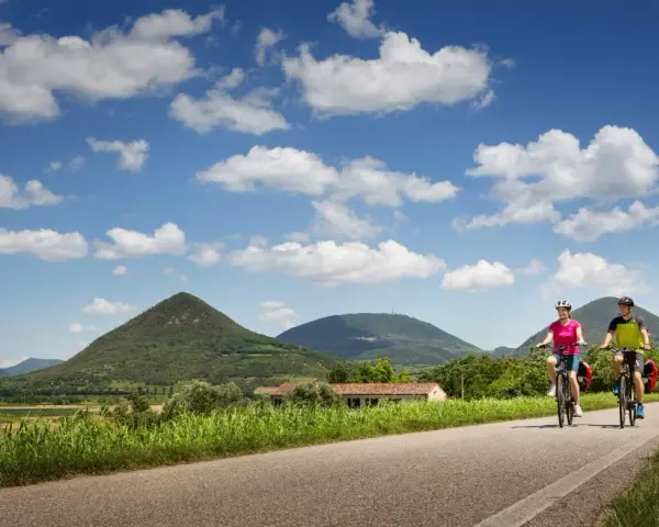 Coppia in bicicletta lungo la ciclabile dei Colli Euganei, tra verdi colline e cielo azzurro con nuvole bianche.