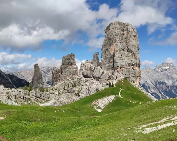 Sentiero che serpeggia tra verdi prati verso le maestose Cinque Torri Dolomiti, sotto un cielo azzurro con nuvole bianche.