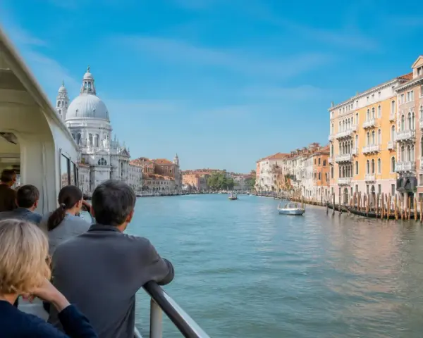 Gente sul vaporetto ammira il Canal Grande, con palazzi colorati e la Basilica di Santa Maria della Salute. Un viaggio tra Murano e Burano.
