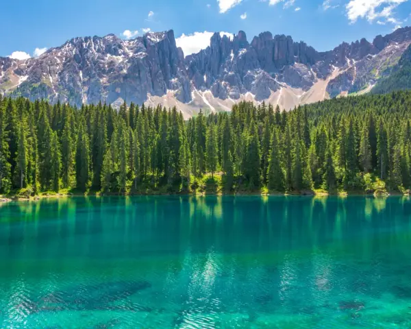 Lago alpino dalle acque turchesi, circondato da una fitta foresta di abeti, con le maestose cime delle Dolomiti innevate sullo sfondo. Un paesaggio da sogno, tra Venezia e le Dolomiti.