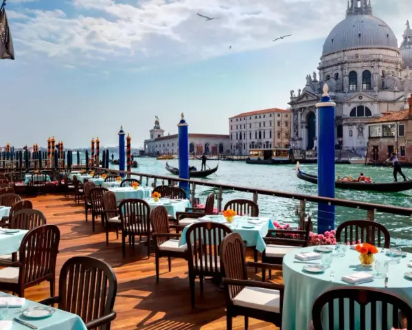 Tavoli apparecchiati con tovaglie azzurre e fiori arancioni, affacciati sul Canal Grande, con gondole e la Basilica di Santa Maria della Salute. Un'ambientazione da sogno per i migliori ristoranti di Venezia.