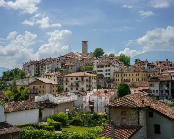 Panoramica di Feltre e dintorni, con case dai tetti rossi che si arrampicano sulla collina sotto un cielo azzurro e nuvoloso.