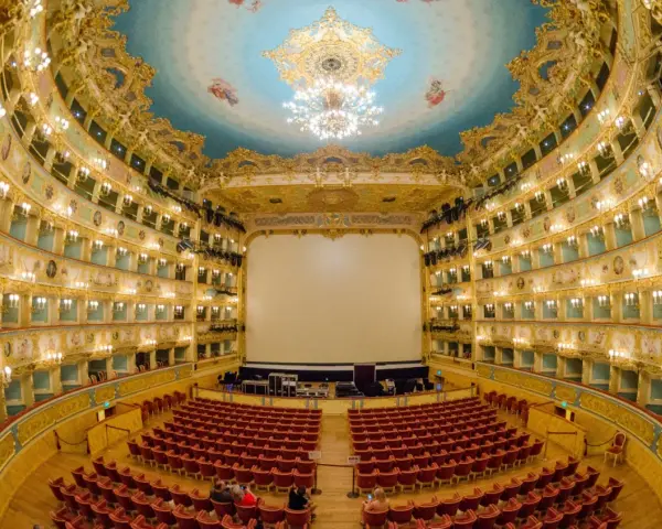 Interno sfarzoso del Teatro La Fenice di Venezia, con palchi dorati, soffitto affrescato e un grande schermo bianco.