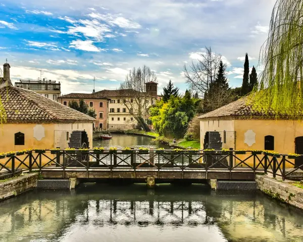 Ponte in legno su un canale con mulini ad acqua, case storiche e vegetazione rigogliosa, un'atmosfera serena che ricorda Portogruaro Caorle.