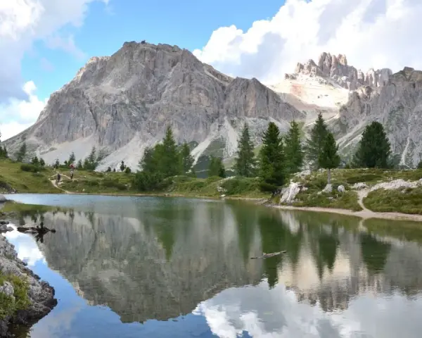 Riflessi perfetti delle montagne nel **lago di Limedes**, con sentiero e pini che incorniciano il paesaggio.