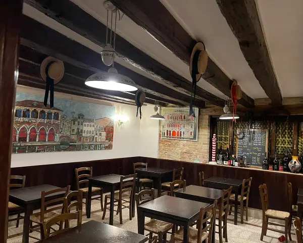 Interno di un **ristorante antico** con dipinti di **Venezia**, tavoli in legno e un calice di vino.