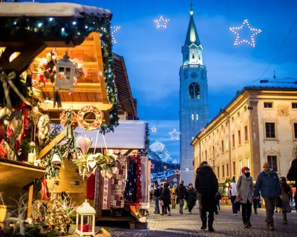 Mercatino di Natale a Cortina d'Ampezzo, con bancarelle addobbate, luci scintillanti e la torre del campanile che domina il cielo serale.