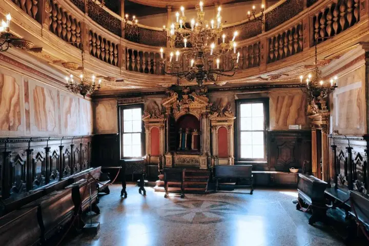 Interno sontuoso della sinagoga di Venezia, con arredi in legno scuro, un grande lampadario e l'Arca Santa dorata.
